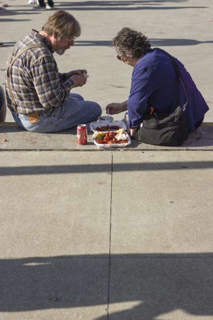 An older man and woman sit along the curb at Clinton Square, a plate of crawfish between them to share.