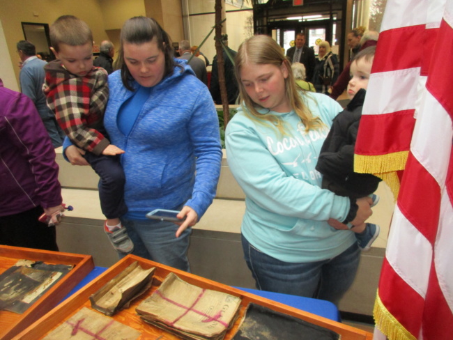 Two women hold toddlers as they look down at a display case. Inside, old books and papers from a recently recovered time capsule give a glimpse to the past.