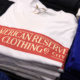 A stack of folded white shirts with a reg "American Reserve Clothing" logo across the chest.