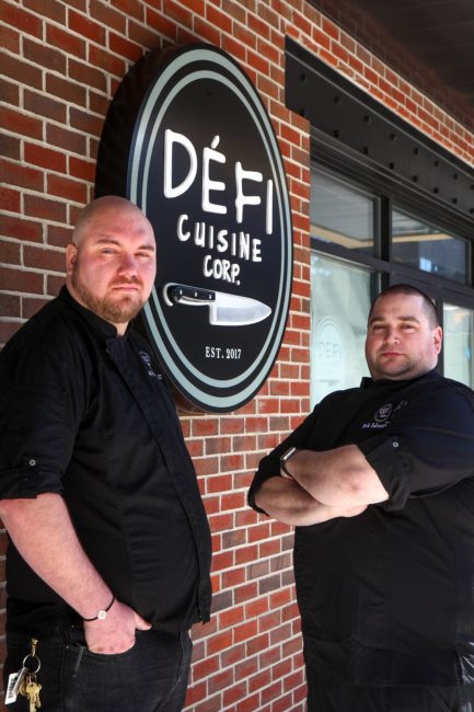 Chefs Cody Dedischew, left, and Nichlas Salvetti, right, stand outside the restaurant in front of the shop's circular, black logo sign.