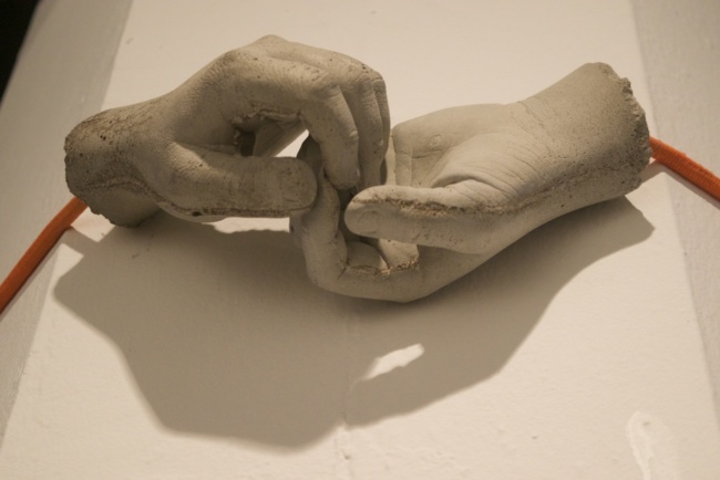 A white, plaster sculpture on a table of two disembodied hands holding at the fingers