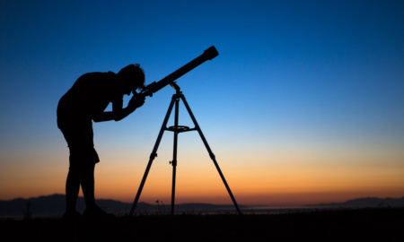 A silhouette of a boy on a clear night looking through a telescope.