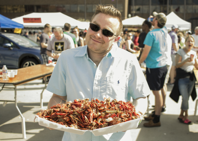 A man in sunglasses holds a massive tray in two hands, piled high with cooked crawfish.