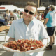 A man in sunglasses holds a massive tray in two hands, piled high with cooked crawfish.
