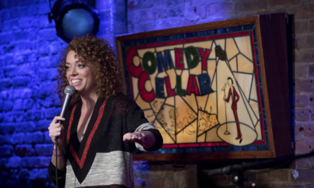 Comedian Michelle Wolf holds a microphone and does a comedy sketch on a stage.