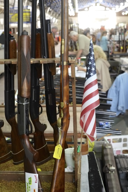 Long guns are propped upright in stands on a table for viewing and purchasing. Each one has a small yellow price tag attached.