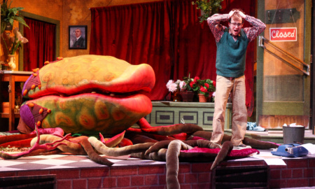 A theater set to look like a diner. To the left is a massive venus flytrap looking plant, bigger than a person. To the right, a man in a sweatervest holds his hands exasperatedly over his heads. Vines tangle across the floor.