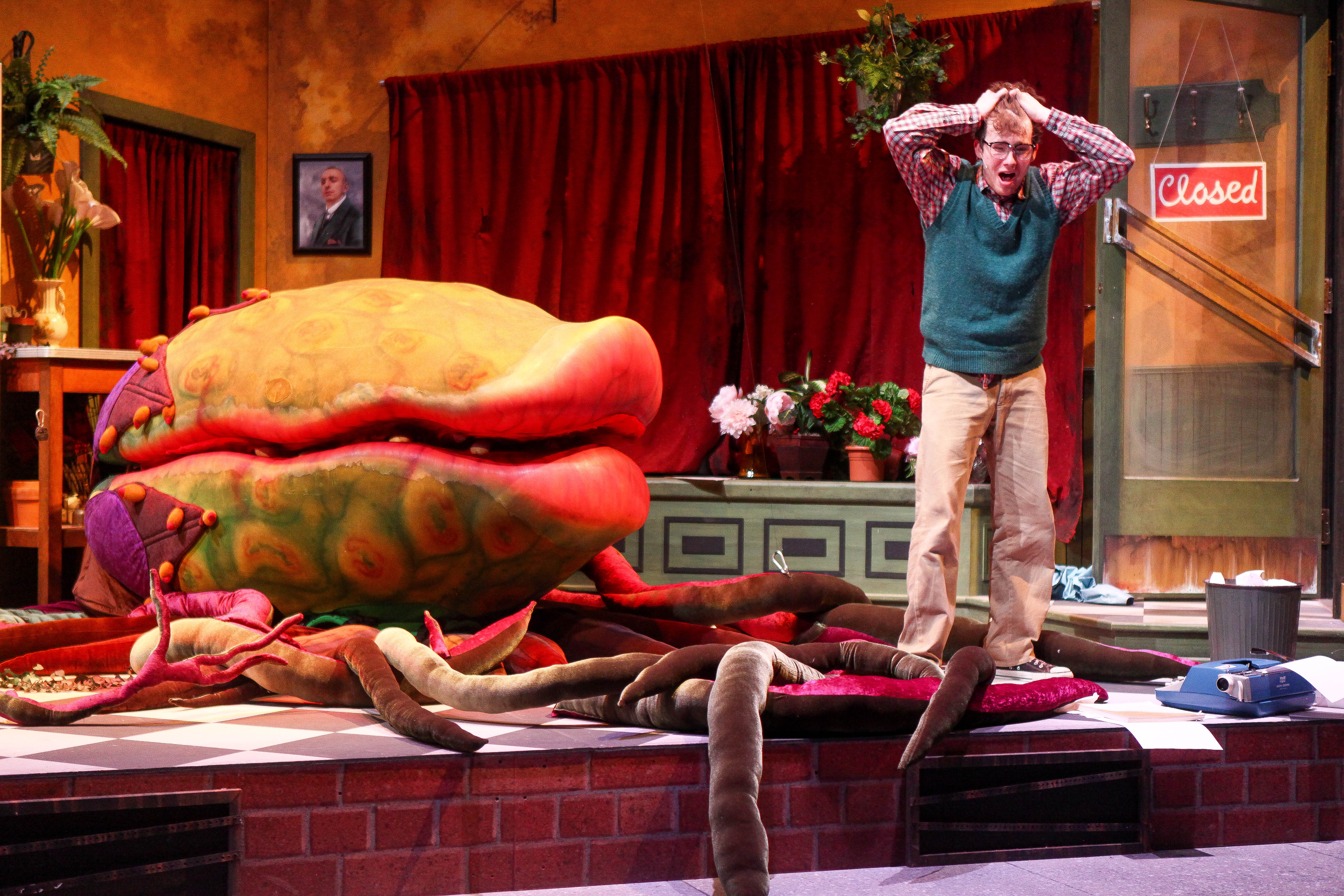 A theater set to look like a diner. To the left is a massive venus flytrap looking plant, bigger than a person. To the right, a man in a sweatervest holds his hands exasperatedly over his heads. Vines tangle across the floor.