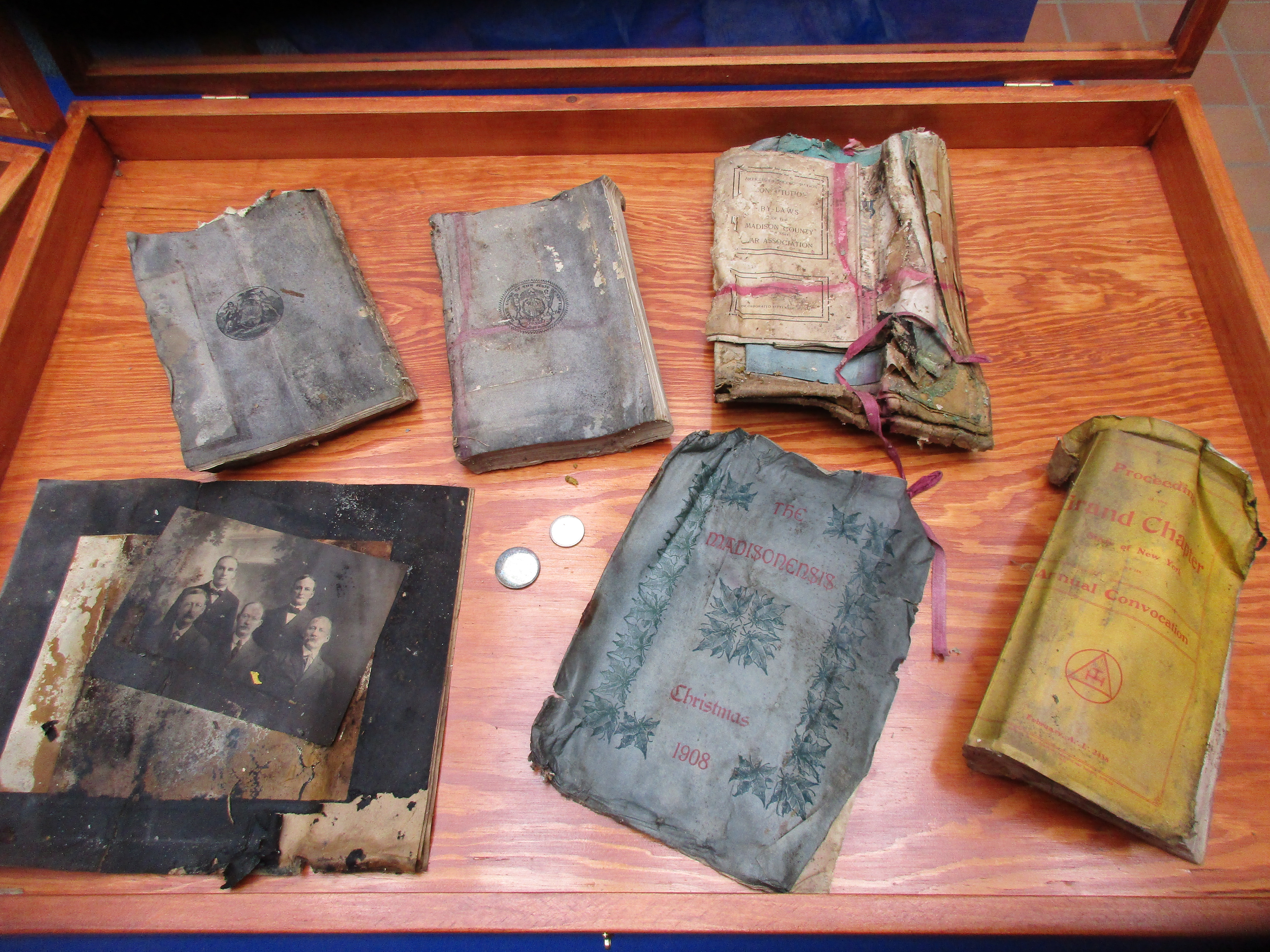 Six old, warped and water damaged books sit in a display case, along with two old coins and some old photos.