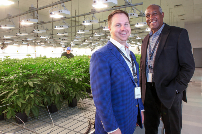 Two men stand off to the right smiling for a photo. Behind them are rows of marijuana plants being grown in a medical facility.