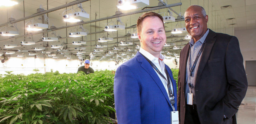 Two men stand off to the right smiling for a photo. Behind them are rows of marijuana plants being grown in a medical facility.