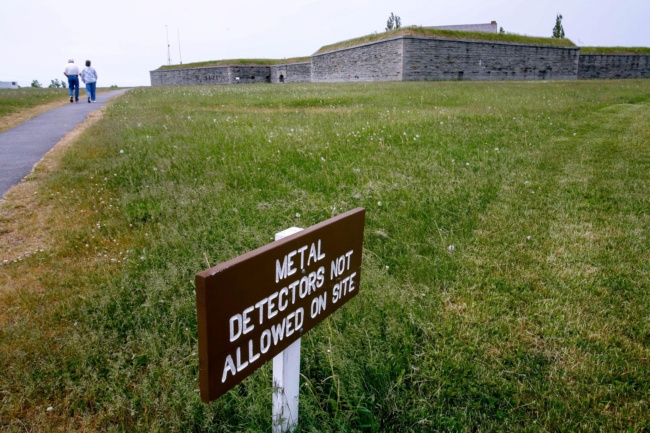 A fort sits at the top of a long, green lawn. A sign in the foreground reads "Metal detectors not allowed on site."