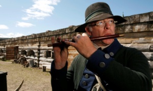 A man dressed as an 18th-century soldier plays a small flute while standing inside a wooden fort during a historical reenactment.