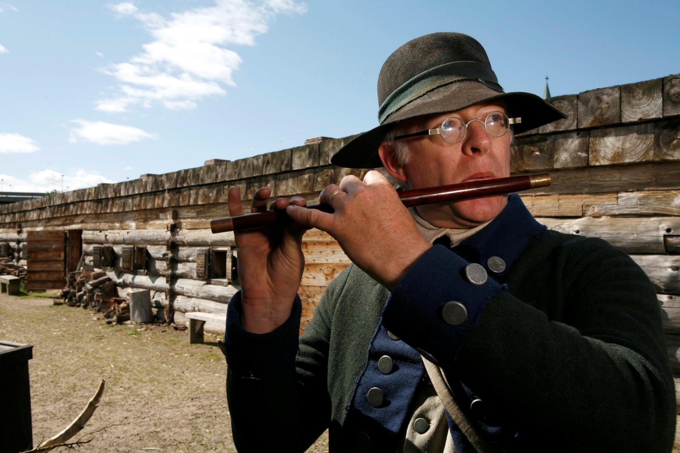 A man dressed as an 18th-century soldier plays a small flute while standing inside a wooden fort during a historical reenactment.