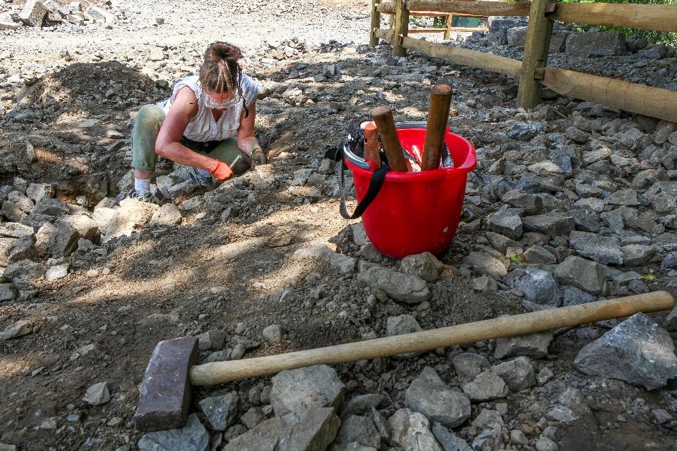 A woman is kneeling on the rocky round, next to a red pail, searching the stones for loose quartz at the Herkimer Diamond Mines.