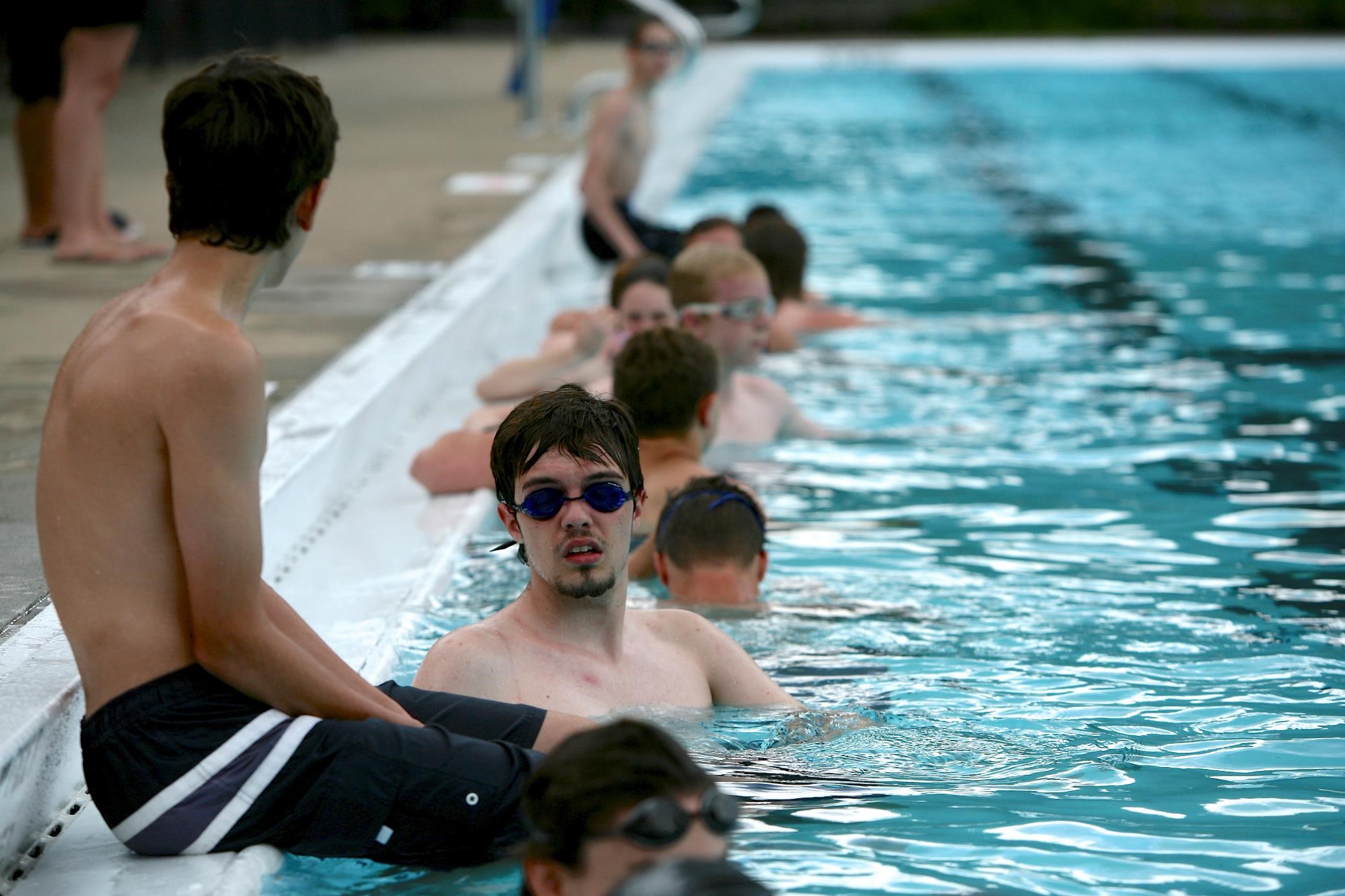 Teens line the side of a pool in the water, wearing goggles, training to be lifeguards.