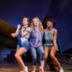 Three young actresses stand in a group, smiling excitedly with their arms around each other, from a scene in Mamma Mia!