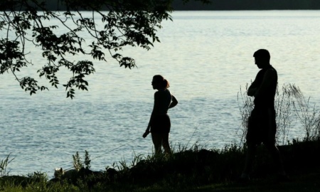 A man and a woman are silhouetted against the waters of Onondaga Lake as they stand at the shore. A small outline of a dog can just be seen past their feet in the tall grass.