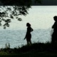 A man and a woman are silhouetted against the waters of Onondaga Lake as they stand at the shore. A small outline of a dog can just be seen past their feet in the tall grass.