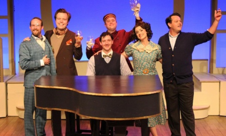 The cast of "Rough Crossing" stand toasting around a piano, dressed in 1930s attire.