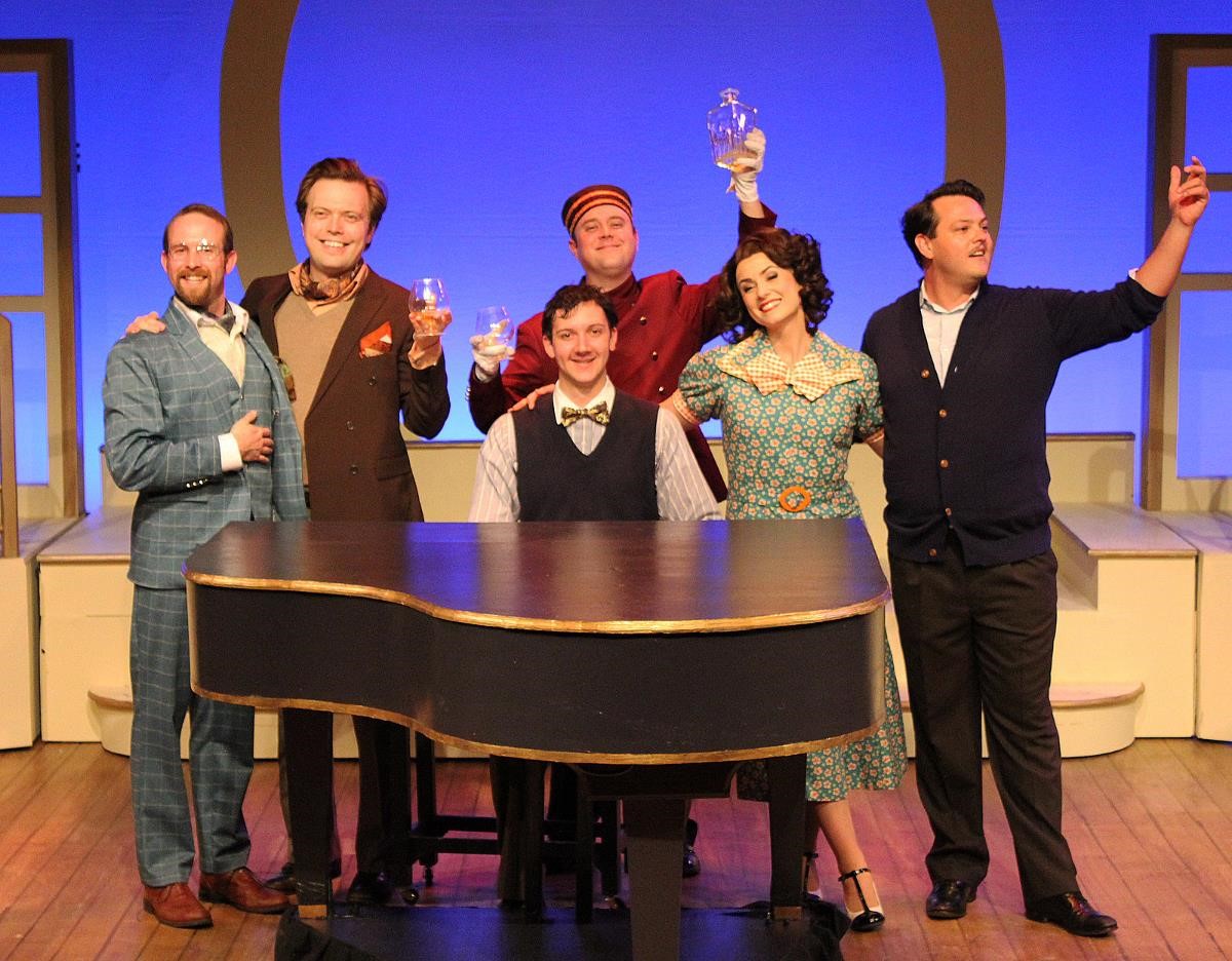 The cast of "Rough Crossing" stand toasting around a piano, dressed in 1930s attire.