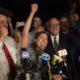Alessandra Biaggi stands in front of several media microphones with a fist in the air as she cheers about her primary win. A large group of aids and supporters crowd around behind her.
