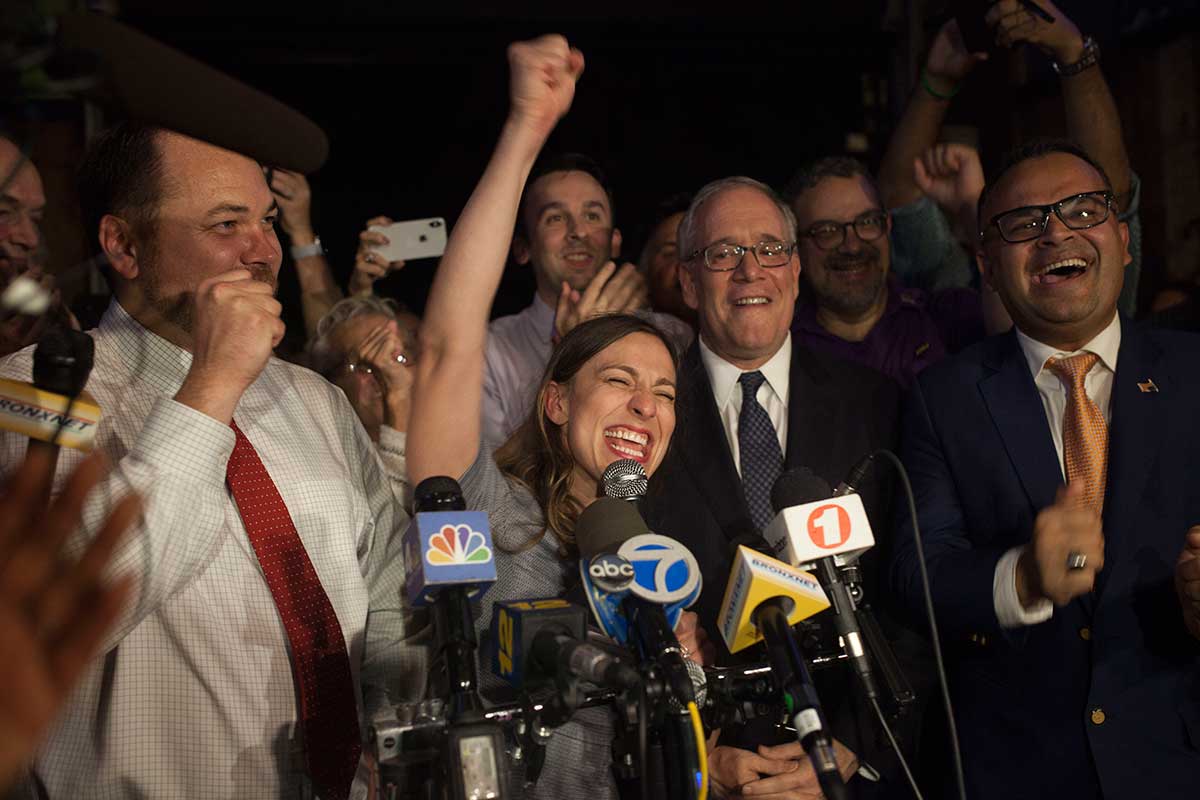 Alessandra Biaggi stands in front of several media microphones with a fist in the air as she cheers about her primary win. A large group of aids and supporters crowd around behind her.